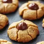Peanut butter cookie blossoms - the image shows a group of freshly baked cookies on a black surface. the cookies are golden brown in color and have a crumbly texture. they are arranged in a scattered manner, with some overlapping each other. in the center of the image, there is a single chocolate chip cookie with a bite taken out of it. the chocolate chip is dark brown and appears to be coated in a thick layer of chocolate. the background is blurred, but it seems to be a kitchen countertop.