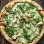 Pesto chicken arugula pizza - the image is of a round pizza on a wooden cutting board. the pizza has a golden brown crust and is topped with a generous amount of melted cheese. on top of the cheese, there are several pieces of grilled chicken arranged in a circular pattern. the chicken appears to be seasoned with herbs and spices, and there are a few sprigs of fresh arugula scattered around the pizza. the background is a dark grey countertop.