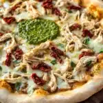 Pesto chicken pizza - the image is of a freshly baked pizza on a wooden cutting board. the pizza has a golden brown crust and is topped with a generous amount of melted cheese. on top of the cheese, there are chunks of chicken, red sun-dried tomatoes, and a green pesto sauce. the chicken appears to be seasoned with herbs and spices, and there are also small pieces of broccoli scattered throughout the pizza. the background is dark, making the pizza stand out.