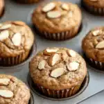 Protein muffin with almond flour - the image shows a muffin tin filled with freshly baked muffins. the muffins are golden brown in color and have a crumbly texture. they are topped with sliced almonds, giving them a crunchy appearance. the tin is sitting on a white marble countertop.