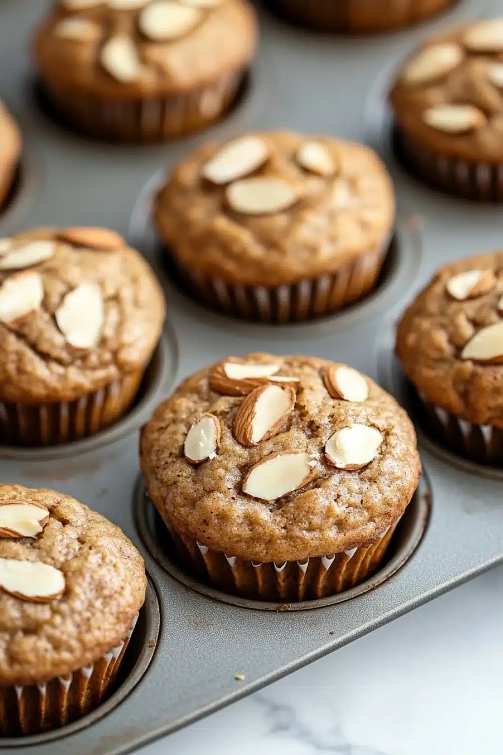 Protein muffin with almond flour - the image shows a muffin tin filled with freshly baked muffins. the muffins are golden brown in color and have a crumbly texture. they are topped with sliced almonds, giving them a crunchy appearance. the tin is sitting on a white marble countertop.