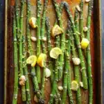 Roasted lemon honey garlic asparagus - the image shows a baking tray with cooked asparagus spears on it. the tray is rectangular in shape and is placed on a wooden table. the spears are arranged in a single layer on the tray, with some of them overlapping each other. on top of the spears, there are small pieces of lemon wedges and garlic cloves scattered around. the garlic cloves are golden brown and appear to be seasoned with herbs and spices. the overall appearance of the dish is fresh and appetizing.