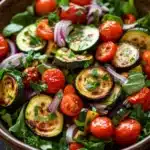 Roasted vegetable salad - the image is a close-up of a colorful salad in a wooden bowl. the salad is made up of various types of vegetables, including zucchini, cherry tomatoes, red onions, and green leafy greens. the vegetables are arranged in a circular pattern, with some overlapping each other. the colors of the vegetables are vibrant and the dish looks fresh and appetizing. the bowl is sitting on a wooden table, and there are a few more cherry tomatoes in the background. the overall mood of the image is fresh and vibrant.
