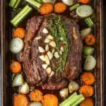 Rosemary garlic roast beef slow cooker - the image shows a large rectangular baking dish filled with a dish of cooked meat and vegetables. the meat appears to be a medium-rare ribeye steak, with a dark brown crust on top. it is garnished with a sprig of rosemary and sliced onions. the vegetables in the dish include carrots, celery, onions, and celery stalks. the dish is sitting on a wooden table with a blue napkin on the side. the overall color scheme of the dish is warm and inviting.