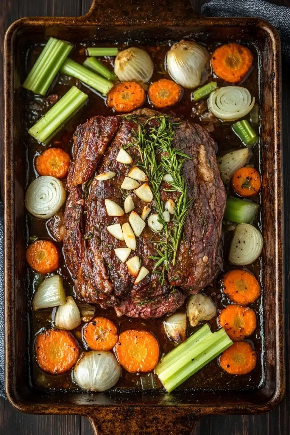 Rosemary garlic roast beef slow cooker - the image shows a large rectangular baking dish filled with a dish of cooked meat and vegetables. the meat appears to be a medium-rare ribeye steak, with a dark brown crust on top. it is garnished with a sprig of rosemary and sliced onions. the vegetables in the dish include carrots, celery, onions, and celery stalks. the dish is sitting on a wooden table with a blue napkin on the side. the overall color scheme of the dish is warm and inviting.