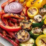 Sauteed vegetables on grill - the image is a close-up of a plate of grilled vegetables. the plate is white with a speckled pattern and is placed on a wooden table. the vegetables are arranged in a circular pattern on the plate. on the left side of the plate, there are sliced red and yellow bell peppers, zucchini, and mushrooms. the bell peppers are grilled to a golden brown color and have a crispy texture. the mushrooms are sliced into thin slices and are placed on top of the vegetables, adding a pop of color to the dish. the overall appearance is colorful and appetizing.