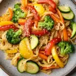 Sauteed vegetables with noodles - the image is a close-up of a plate of noodles with various vegetables. the noodles are light brown in color and appear to be stir-fry. the vegetables include broccoli, red bell peppers, yellow zucchini, and red bell pepper. there are also sesame seeds sprinkled on top of the noodles. the plate is sitting on a white marble countertop.