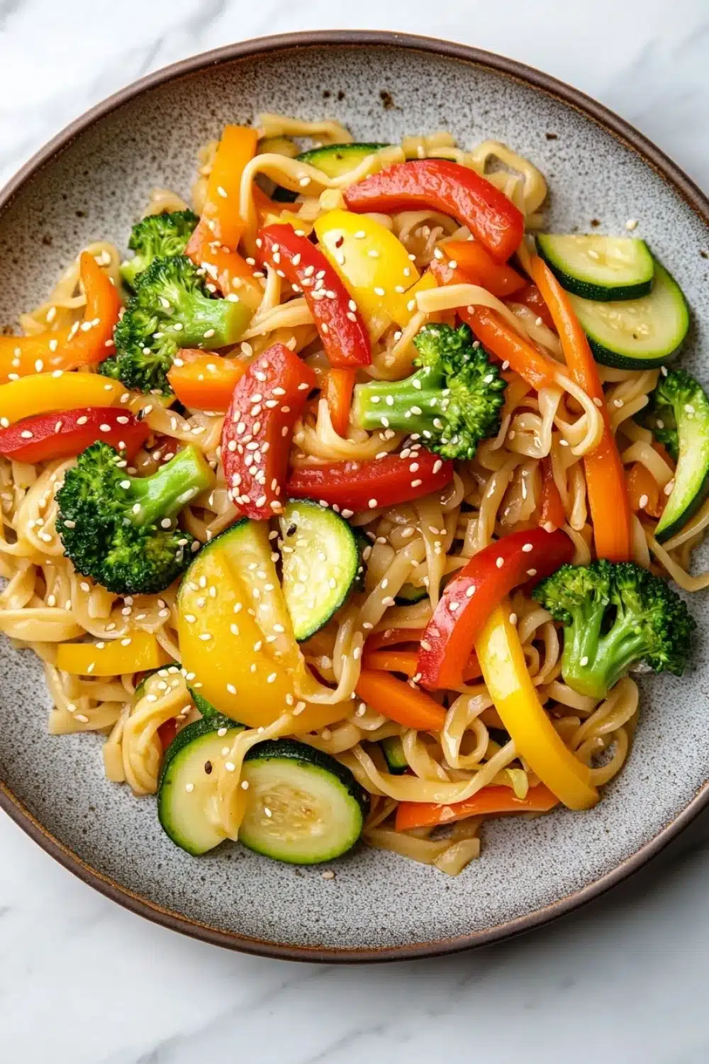 Sauteed vegetables with noodles - the image is a close-up of a plate of noodles with various vegetables. the noodles are light brown in color and appear to be stir-fry. the vegetables include broccoli, red bell peppers, yellow zucchini, and red bell pepper. there are also sesame seeds sprinkled on top of the noodles. the plate is sitting on a white marble countertop.