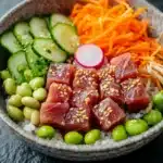 Seared tuna poke bowl - the image is a close-up of a bowl of food. the bowl is made of dark brown ceramic and is placed on a dark grey textured surface. the food in the bowl appears to be a poke bowl with a variety of ingredients. on top of the poke bowl, there are sliced cucumbers, carrots, and edamame, as well as sliced radishes and sesame seeds. the rice is white and fluffy, and the food is arranged in an aesthetically pleasing manner. the colors of the ingredients are vibrant and the dish looks fresh and appetizing.