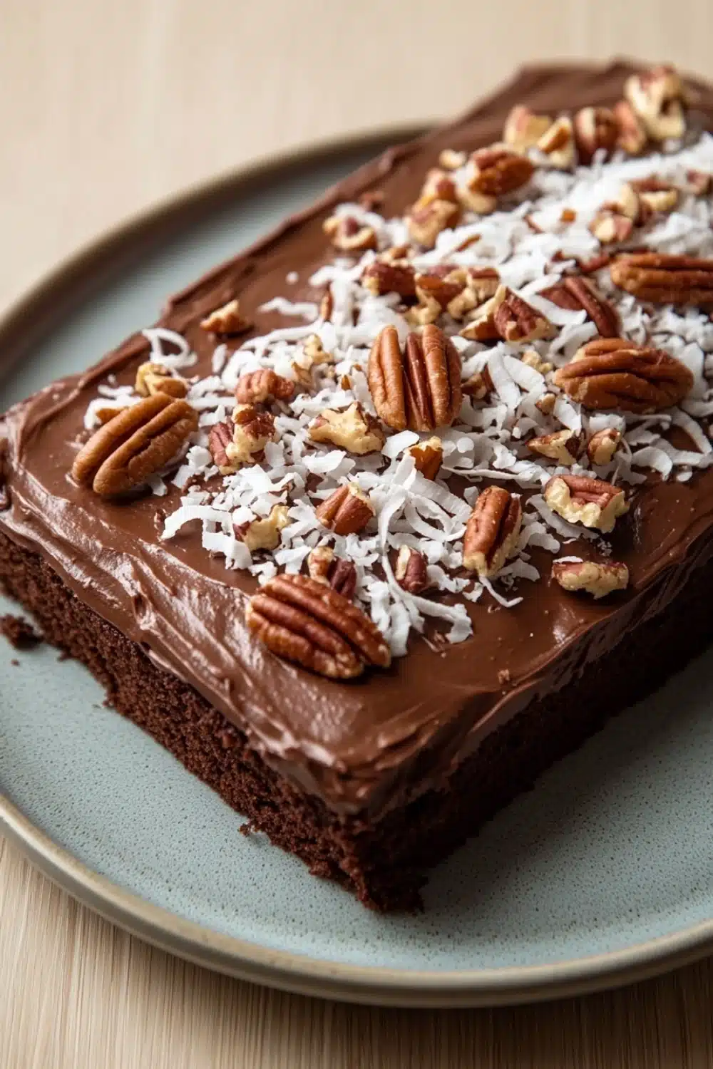 Sheet cake german chocolate - the image is of a slice of chocolate cake on a blue plate. the cake has a thick layer of chocolate frosting on top, with chopped pecans scattered throughout. the frosting is a rich, dark brown color and appears to be smooth and glossy. the pecan pieces are arranged in a scattered manner, with some overlapping each other. the plate is sitting on a wooden table, and the background is blurred, making the cake the focal point of the image.