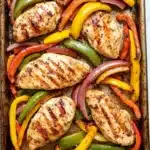 Sheet pan chicken breast with fajita veggies - the image shows a rectangular baking tray filled with grilled chicken and colorful bell peppers. the chicken is cooked to a golden brown color and appears to be seasoned with herbs and spices. the bell peppers are of different colors - red, yellow, green, and orange - and are arranged in a circular pattern around the chicken. the tray is lined with parchment paper and is sitting on a white marble countertop.