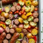 Sheet pan dinner sausage and potatoes - the image shows a baking tray filled with a variety of ingredients. on the left side of the tray, there are two sausages, one red and one yellow, which appear to be cooked sausage. next to the sausage, there is a pile of diced potatoes, red onions, and yellow bell peppers. the potatoes are cut into small cubes and are arranged in a pile on top of each other. the tray is lined with parchment paper and is garnished with fresh parsley. the background is a white marble countertop.
