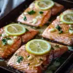 Sheet pan garlic lemon butter salmon - the image shows four pieces of salmon on a baking tray. the salmon is cooked to a golden brown color and appears to be seasoned with herbs and lemon wedges. the tray is lined with parchment paper and there are a few sprigs of parsley scattered around the tray. in the background, there are more lemons and a white napkin. the overall aesthetic of the image is rustic and appetizing.