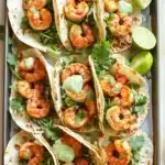 Sheet pan spicy shrimp tacos with jalapeno lime crema - the image shows a tray of six shrimp tacos on a wooden table. the tacos are made with tortilla shells and are filled with shrimp, avocado, and cilantro. there are also two lime wedges on the tray. the tray is lined with parchment paper and there are a few sprigs of cilantro scattered around the tray, adding a pop of green color to the dish. the background is blurred, but it appears to be a kitchen countertop.