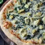 Sheet pan spinach dip pizza - the image is of a freshly baked pizza on a wooden cutting board. the pizza has a golden brown crust and is topped with melted cheese and artichokes. the cheese is melted and bubbly, and there are small pieces of spinach scattered throughout the pizza. the spinach is a vibrant green color and appears to be wilted and wilted. there is a sprig of fresh basil on the side of the cutting board, adding a pop of color to the dish. the background is dark and out of focus, making the pizza the focal point of the image.