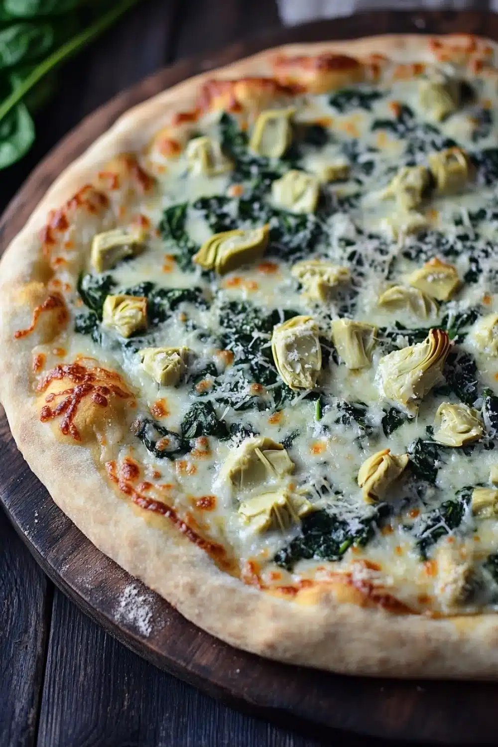 Sheet pan spinach dip pizza - the image is of a freshly baked pizza on a wooden cutting board. the pizza has a golden brown crust and is topped with melted cheese and artichokes. the cheese is melted and bubbly, and there are small pieces of spinach scattered throughout the pizza. the spinach is a vibrant green color and appears to be wilted and wilted. there is a sprig of fresh basil on the side of the cutting board, adding a pop of color to the dish. the background is dark and out of focus, making the pizza the focal point of the image.