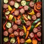 Sheet pan vegetables and sausage - the image shows a rectangular baking tray filled with a variety of colorful vegetables. the vegetables include sliced zucchini, carrots, bell peppers, and onions. the tray is lined with parchment paper and is placed on a wooden table. the colors of the vegetables are vibrant and the dish looks appetizing. there are also some herbs and spices scattered throughout the tray, adding a pop of green to the dish.