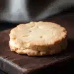 Shortbread cookie illustration - the image shows a single cookie on a wooden cutting board. the cookie appears to be freshly baked and has a golden brown color. it has a crumbly texture and is slightly uneven on the edges. the cutting board is dark and has some scratches and marks on it. the background is blurred, but it seems to be a kitchen countertop with a white cloth napkin on the right side. the overall mood of the image is rustic and cozy.