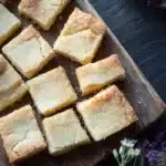 Shortbread cookie squares - the image shows a wooden cutting board with several square-shaped pieces of cake arranged in a grid-like pattern. the cake appears to be freshly baked and has a golden-brown crust on top. the board is placed on a dark wooden table with a few purple flowers scattered around it. the background is blurred, but it seems to be a kitchen countertop. the overall mood of the image is rustic and cozy.
