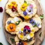 Shortbread cookie with edible flowers - the image shows a wooden plate with six freshly baked cookies arranged in a circular pattern. the cookies are golden brown and have a crumbly texture. each cookie is decorated with colorful pansies in shades of yellow, orange, purple, and white. the pansies are scattered around the plate, adding a pop of color to the image. the plate is surrounded by a few green leaves, giving it a natural and rustic feel. the background is a white marble surface.