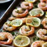 Shrimp sheet pan lemon - the image shows a baking tray with cooked shrimp on it. the shrimp are pink and appear to be seasoned with herbs and lemon wedges. the tray is lined with parchment paper and there are a few sprigs of parsley scattered around the tray. in the background, there is a white plate with a fork and knife on it, suggesting that the shrimp are ready to be served. the overall color scheme of the image is warm and inviting.