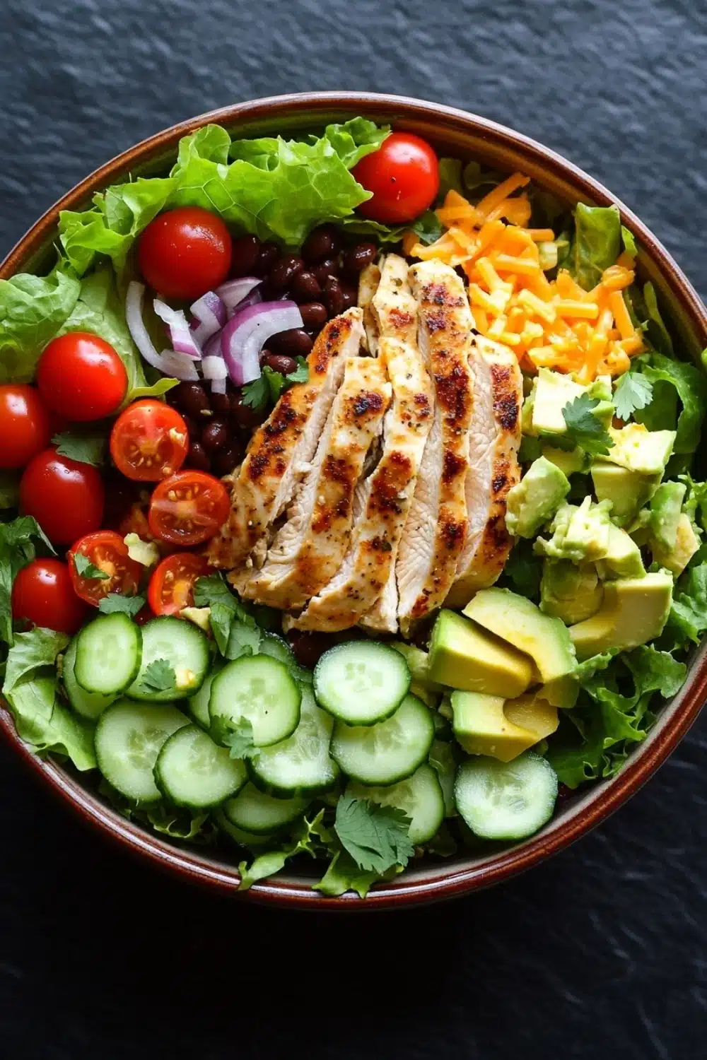 Siracha lime chicken chopped salad - the image is of a large bowl filled with a colorful salad. the salad is made up of various greens, including lettuce, cherry tomatoes, black beans, and sliced avocado. on top of the salad, there is a piece of grilled chicken that appears to be seasoned with herbs and spices. the chicken is golden brown and looks crispy on the outside. the bowl is sitting on a black countertop.