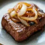Slow cooker beef cube steak - the image shows a plate of food with a piece of steak on it. the steak appears to be cooked medium-rare and has a dark brown crust on the outside. on top of the steak, there are sliced onions arranged in a circular pattern. the onions are golden brown and appear to be seasoned with herbs and spices. the plate is a light blue color with a speckled pattern around the edges. the background is blurred, but it seems to be a wooden table.