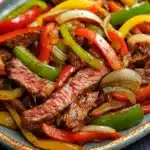 Slow cooker beef fajitas slow cooker - the image is a close-up of a plate of food. the plate is made of ceramic and has a blue rim. the food appears to be a stir-fry with sliced red, yellow, and green bell peppers, onions, and beef. the beef is cooked medium-rare and is arranged in a pile on top of the vegetables. the colors of the ingredients are vibrant and the dish looks appetizing. the background is dark and out of focus, making the plate and vegetables stand out.