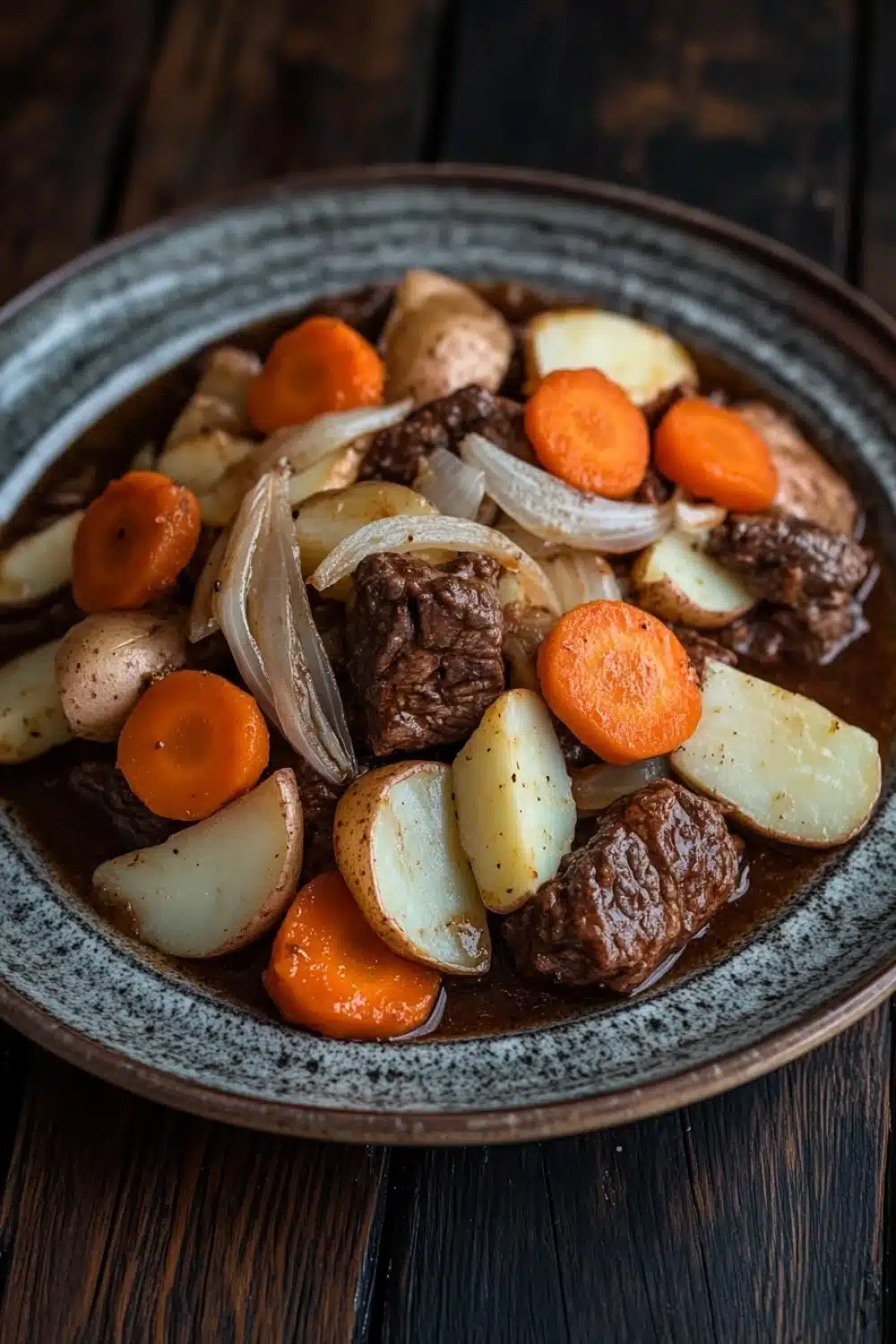 Slow cooker beef hotpot - the image is a close-up of a bowl of beef stew on a wooden table. the stew is made with chunks of beef, potatoes, carrots, onions, and other vegetables. the beef is cooked in a rich, dark brown sauce and appears to be seasoned with herbs and spices. the potatoes are cut into small cubes and are arranged in a pile on top of the beef. the carrots are bright orange and appear to be fresh. the onions are sliced and are scattered throughout the stew. the bowl is made of dark brown ceramic with a speckled pattern around the edges. the background is blurred, making the stew the focal point of the image.