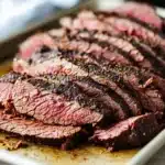 Slow cooker deli roast beef - the image shows a plate of sliced steak on a white rectangular plate. the steak appears to be cooked medium-rare, with a pinkish-red color and a slightly charred texture. the slices are arranged in a single layer on the plate, with some overlapping each other. the plate is sitting on a wooden table, and there is a white napkin on the side. the background is blurred, but it seems to be a kitchen countertop.