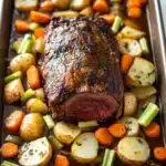 Slow cooker pot roast beef - the image shows a rectangular baking tray filled with a roast beef dish. the roast beef is cooked medium-rare and has a golden brown crust on top. it is garnished with herbs and spices. the dish is made up of diced potatoes, carrots, and celery, all cooked in a rich, dark brown sauce. the potatoes are cut into small cubes and are arranged in a single layer on the tray. the carrots are bright orange and the celery is a vibrant green color. the tray is sitting on a white marble countertop.