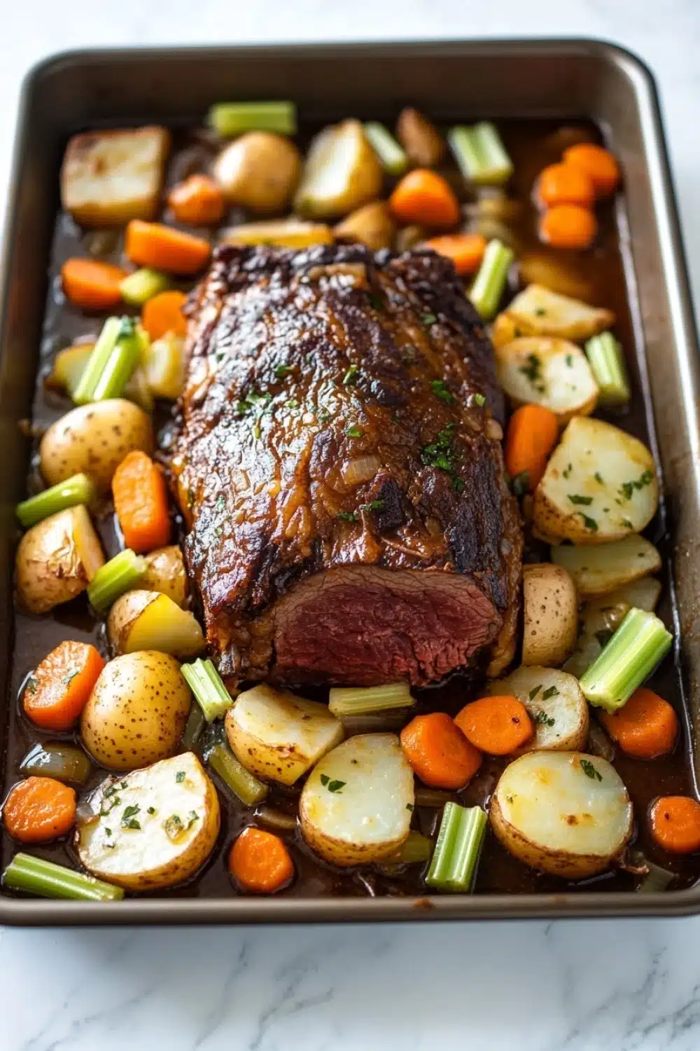 Slow cooker pot roast beef - the image shows a rectangular baking tray filled with a roast beef dish. the roast beef is cooked medium-rare and has a golden brown crust on top. it is garnished with herbs and spices. the dish is made up of diced potatoes, carrots, and celery, all cooked in a rich, dark brown sauce. the potatoes are cut into small cubes and are arranged in a single layer on the tray. the carrots are bright orange and the celery is a vibrant green color. the tray is sitting on a white marble countertop.