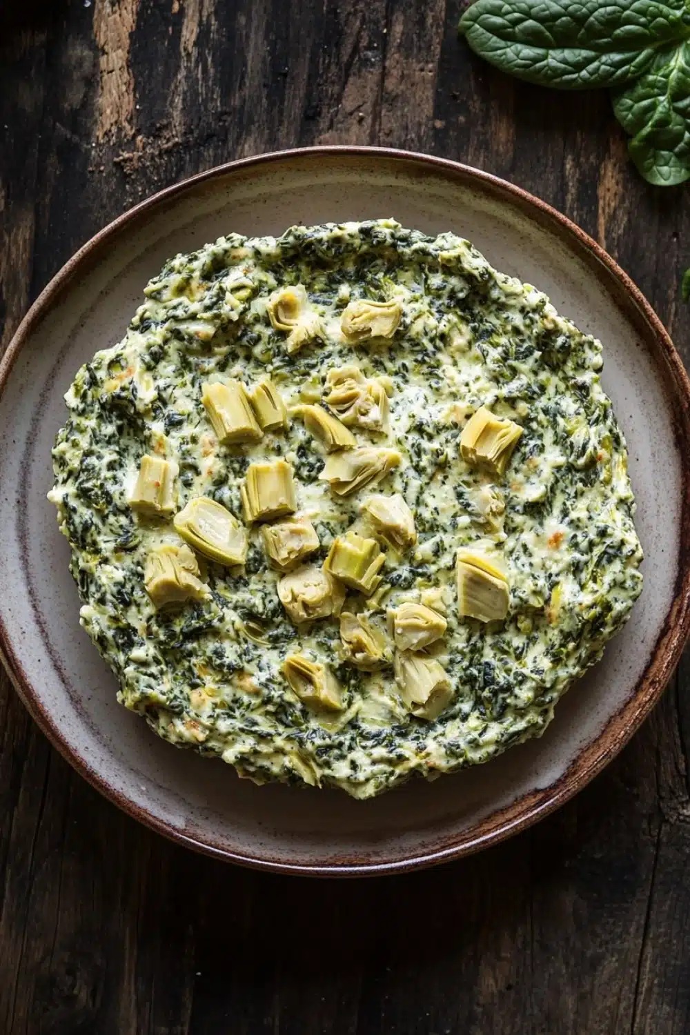 Spinach artichoke dip no cheese - the image is a top view of a round dish on a wooden table. the dish appears to be a type of spinach and artichoke dip, with a creamy green color and small pieces of artichokes scattered throughout. the plate is made of a light-colored wood and has a brown rim. on the right side of the image, there is a sprig of fresh basil leaves, adding a pop of green color to the dish. the background is dark and out of focus, making the dish stand out.