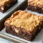 Sugar cookie brownies - the image shows a tray of freshly baked brownies on a white marble countertop. the brownies are rectangular in shape and have a rich, dark brown color. they are topped with a layer of peanut butter frosting, which is a light brown color and has a crumbly texture. the frosting appears to be slightly uneven and has small white chocolate chips scattered throughout. the tray is lined with parchment paper and there are a few more brownies visible in the background.
