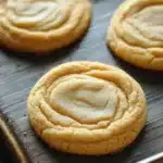Sugar cookie mix - the image shows a baking tray with six freshly baked cookies on it. the cookies are round and golden brown in color, with a swirl pattern on top. they are arranged in a row on the tray, with some overlapping each other. the tray is lined with parchment paper, and the cookies appear to be freshly baked. the background is blurred, but it appears to be a wooden table.