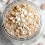 Sugar cookie overnight oats - the image is a close-up of a glass jar filled with a creamy, light-colored dessert. the dessert appears to be oatmeal or oatmeal, with small white marshmallows scattered on top. the jar is sitting on a white marble surface, and there are scattered chocolate chips around the jar. the background is blurred, making the jar the focal point of the image.