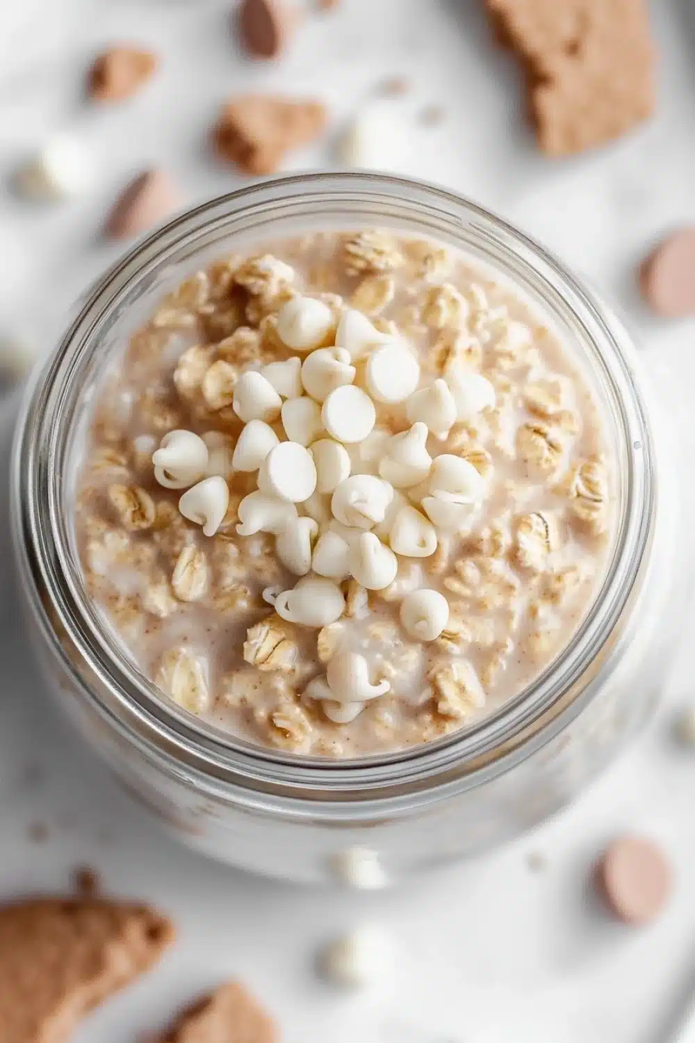 Sugar cookie overnight oats - the image is a close-up of a glass jar filled with a creamy, light-colored dessert. the dessert appears to be oatmeal or oatmeal, with small white marshmallows scattered on top. the jar is sitting on a white marble surface, and there are scattered chocolate chips around the jar. the background is blurred, making the jar the focal point of the image.