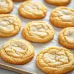 Sugar cookie platter - the image shows a tray of freshly baked cookies on a white countertop. the cookies are round and golden brown in color, with a crumbly texture on top. they are arranged in a single layer on the tray, with some overlapping each other. some of the cookies have a light dusting of powdered sugar on top, indicating that they have been freshly baked. the tray appears to be lined with parchment paper, and the background is blurred, making the cookies the focal point of the image.