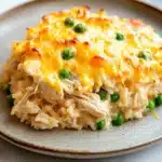 Tiktok chicken and rice casserole - the image shows a plate of food on a white tablecloth. the plate is round and has a light brown color with a speckled pattern. on top of the plate, there is a layer of shredded chicken and peas. the chicken appears to be cooked and is covered in melted cheese. the peas are bright green and look fresh and vibrant. the dish looks appetizing and ready to eat.