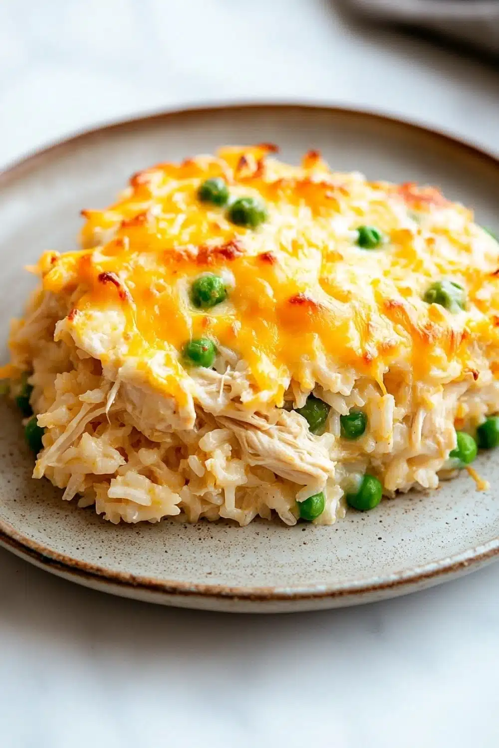 Tiktok chicken and rice casserole - the image shows a plate of food on a white tablecloth. the plate is round and has a light brown color with a speckled pattern. on top of the plate, there is a layer of shredded chicken and peas. the chicken appears to be cooked and is covered in melted cheese. the peas are bright green and look fresh and vibrant. the dish looks appetizing and ready to eat.