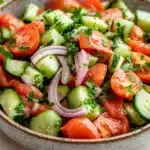 Tomato cucumber and onion salad - the image is a close-up of a bowl of salad. the salad is made up of diced tomatoes, cucumbers, and red onions. the tomatoes are cut into small cubes and are arranged in a circular pattern. the cucumbers are sliced into thin strips and are scattered throughout the salad. there are also some fresh herbs, such as cilantro, scattered throughout. the bowl is sitting on a white tablecloth with a sprig of parsley on the side. the background is blurred, but it appears to be a kitchen countertop.
