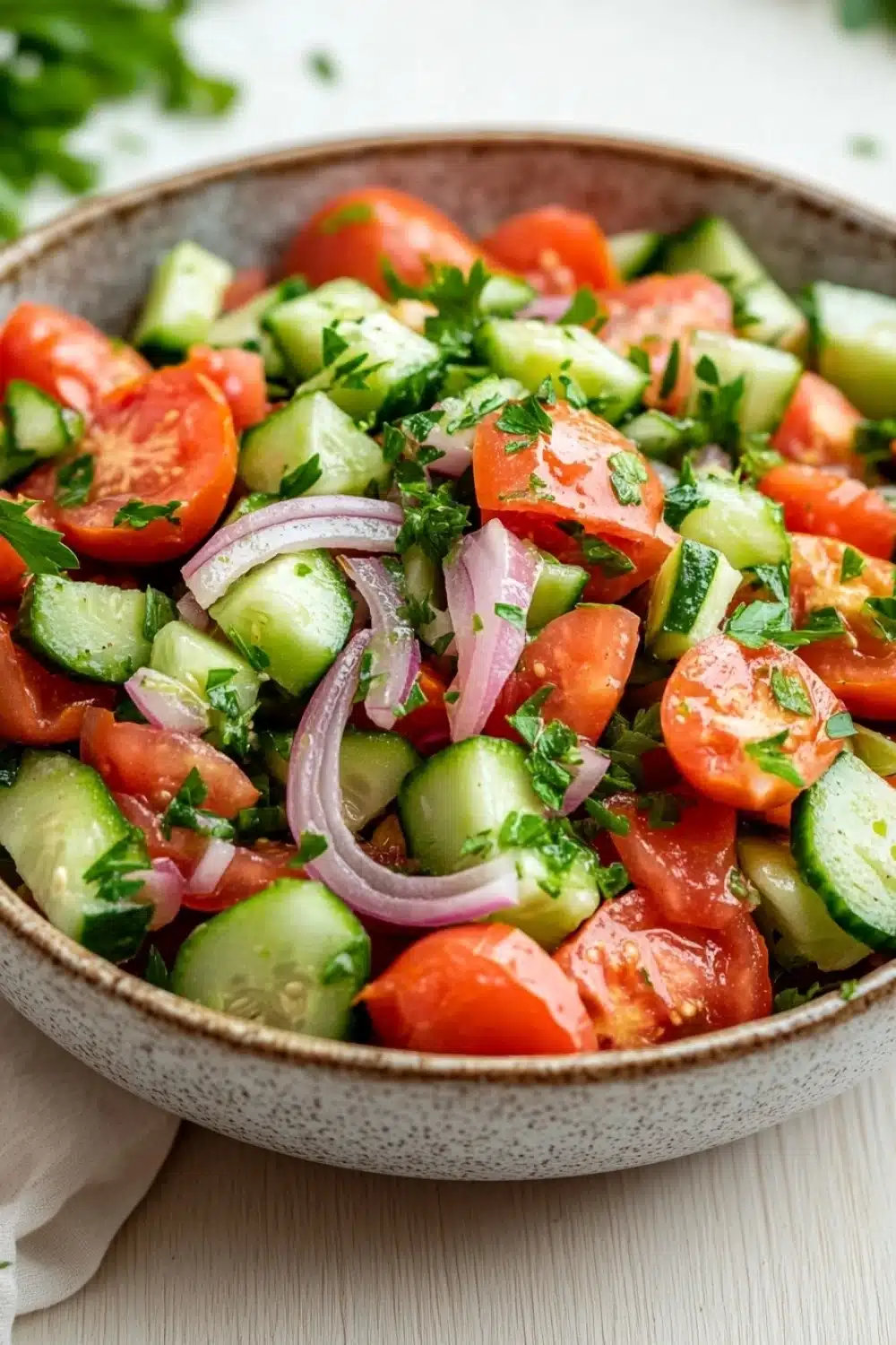 Tomato cucumber and onion salad - the image is a close-up of a bowl of salad. the salad is made up of diced tomatoes, cucumbers, and red onions. the tomatoes are cut into small cubes and are arranged in a circular pattern. the cucumbers are sliced into thin strips and are scattered throughout the salad. there are also some fresh herbs, such as cilantro, scattered throughout. the bowl is sitting on a white tablecloth with a sprig of parsley on the side. the background is blurred, but it appears to be a kitchen countertop.