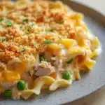 Tuna casserole classic - the image shows a plate of macaroni and cheese casserole. the dish is made up of long, thin noodles that are covered in a creamy yellow sauce. the sauce appears to be melted and bubbly, with chunks of ham and peas scattered throughout. on top of the sauce, there is a layer of breadcrumbs, which are golden brown and crispy. the plate is white and round, and it is sitting on a wooden table.