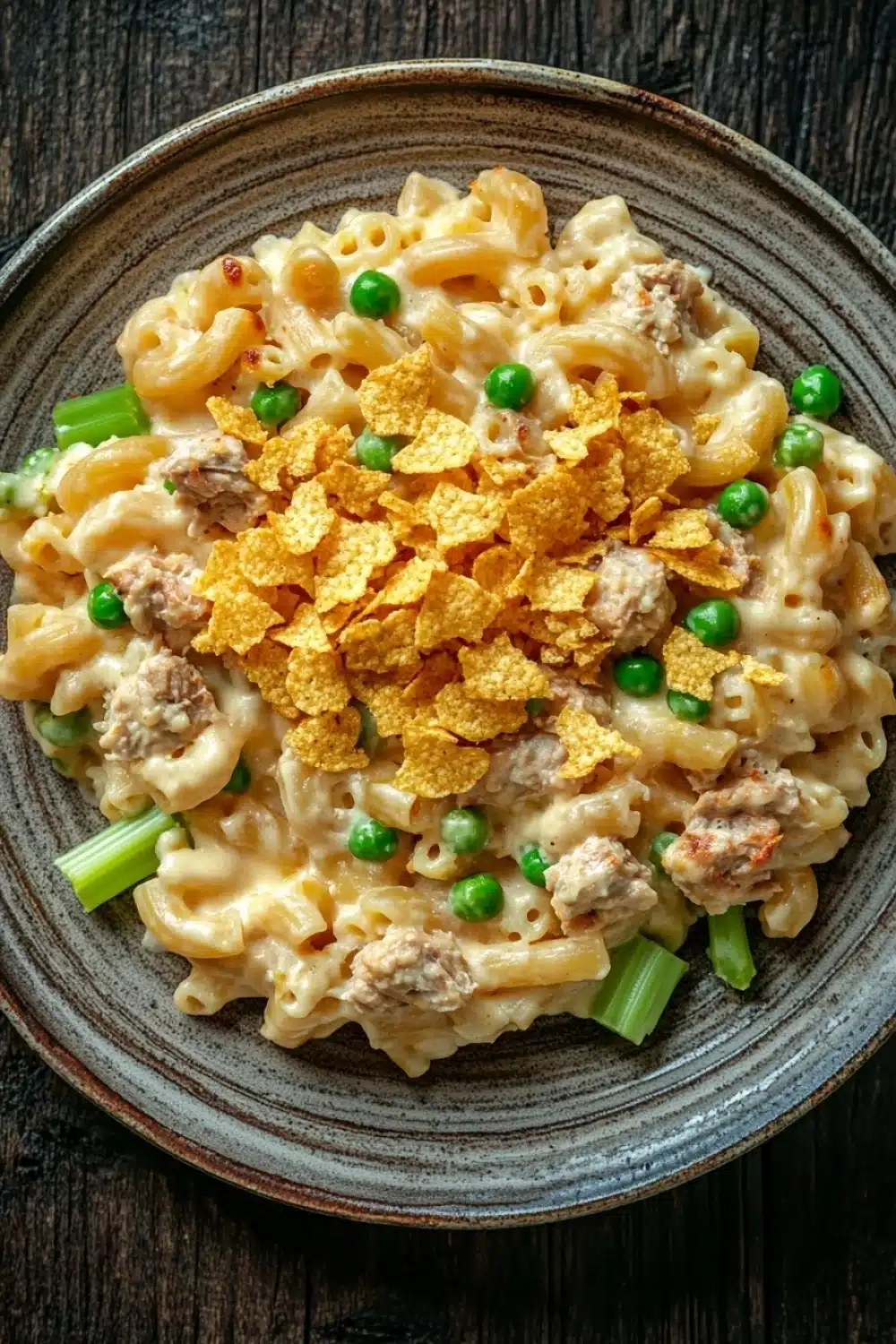Tuna casserole with mac and cheese - the image is a close-up of a bowl of macaroni and cheese. the bowl is made of dark brown ceramic and is placed on a wooden table. the pasta is a creamy yellow color and appears to be cooked al dente. on top of the pasta, there are chunks of green peas and a sprinkle of orange corn flakes. the dish looks appetizing and ready to eat.