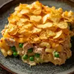 Tuna casserole with potato chips - the image shows a plate of macaroni and cheese casserole on a wooden table. the dish is made with macaronis, cheese, and green peas, and is topped with a layer of melted cheese. there are also a few tortilla chips scattered on top of the dish. the plate is brown and has a speckled pattern around the edges. the background is blurred, but it appears to be a rustic wooden table with a dark finish.