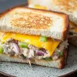 Tuna melt sandwiches - the image shows a close-up of a grilled cheese sandwich on a plate. the sandwich is made with two slices of toasted bread, with melted cheese oozing out of the sides. the cheese is a bright yellow color and appears to be melted and bubbly. on top of the cheese, there are chunks of chicken, red onions, and green bell peppers. the plate is white with a speckled pattern. the background is dark, making the sandwich stand out.