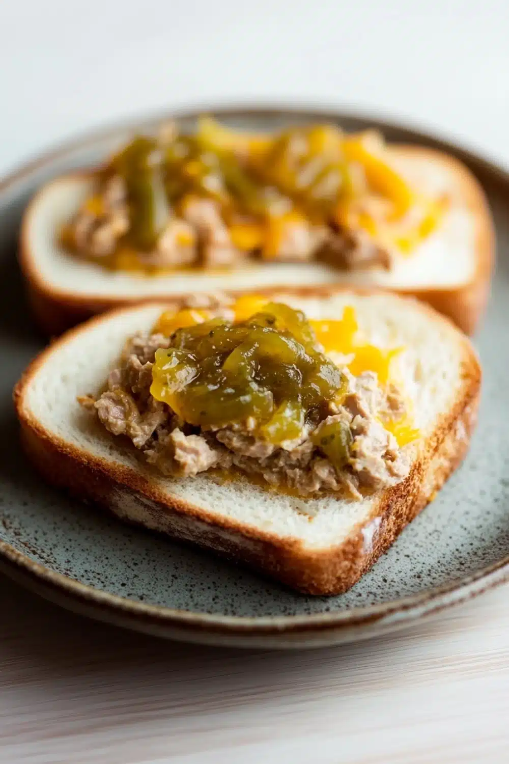 Tuna melt with relish - the image shows two slices of bread on a blue and white plate. the bread appears to be toasted and has a golden brown crust. on top of the bread, there is a mixture of ground meat, possibly tuna or tuna, with a dollop of green sauce on top. the sauce is drizzled over the top, giving it a glossy sheen. the plate is sitting on a white tablecloth.
