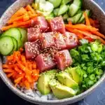 Tuna poke bowl spicy - the image is a close-up of a bowl of food. the bowl is made of a light-colored ceramic material and is placed on a dark grey countertop. inside the bowl, there are various ingredients such as sliced cucumbers, sliced carrots, sliced avocado, and chopped green onions. on top of the vegetables, there is a layer of sesame seeds. the ingredients are arranged in a colorful and appetizing manner.