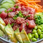 Tuna poke bowl spicy mayo - the image is a close-up of a bowl of food. the bowl is made of ceramic and is sitting on a wooden table. the food in the bowl appears to be a colorful and appetizing dish with a variety of ingredients. the main focus of the image is on a bed of white rice, which is topped with sliced avocado, sliced carrots, and edamame. the avocado is cut into small cubes and is drizzled with sesame seeds. the carrots are bright orange and appear to be freshly cut. the edams are a vibrant green color and are scattered throughout the dish, adding a pop of color to the plate. the rice is fluffy and white, and the vegetables are arranged in a way that creates a contrast with the bright colors of the food.