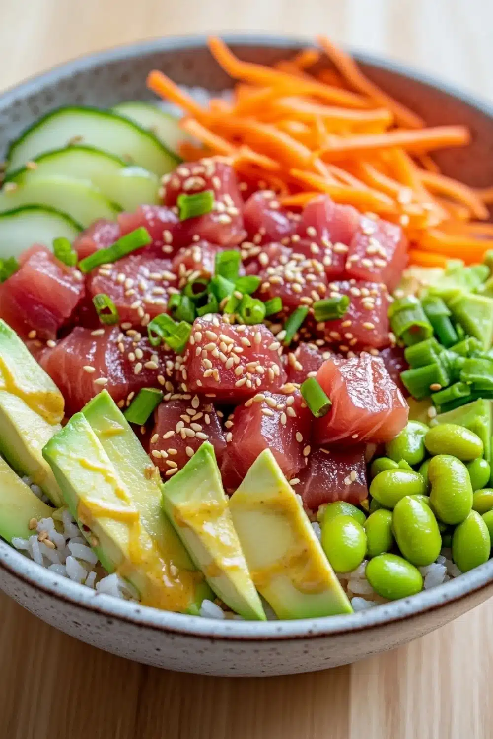 Tuna poke bowl spicy mayo - the image is a close-up of a bowl of food. the bowl is made of ceramic and is sitting on a wooden table. the food in the bowl appears to be a colorful and appetizing dish with a variety of ingredients. the main focus of the image is on a bed of white rice, which is topped with sliced avocado, sliced carrots, and edamame. the avocado is cut into small cubes and is drizzled with sesame seeds. the carrots are bright orange and appear to be freshly cut. the edams are a vibrant green color and are scattered throughout the dish, adding a pop of color to the plate. the rice is fluffy and white, and the vegetables are arranged in a way that creates a contrast with the bright colors of the food.